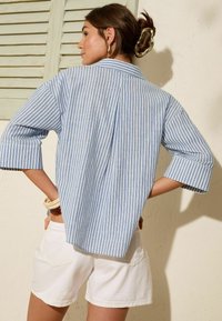 Camicia a righe blu e bianche con vestibilità ampia e maniche arrotolate a tre quarti, abbinata a pantaloni corti bianchi, davanti a un muro texture.
