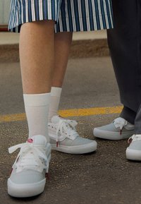 Weiße Sneakers mit grauen Akzenten, ausgestattet mit Schnürsenkeln und durchlässigen Details. Tragen mit weißen Socken, kombiniert mit einem gestreiften Rock und grauen Hosen.
