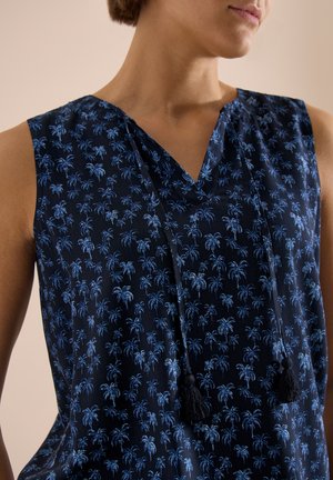 Top sans manches bleu marine avec motif de palmier bleu, encolure en V et deux liens pompons suspendus sur une personne aux cheveux courts, fond beige.