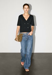 Schwarzes, kurzärmeliges, bauchfreies Oberteil mit V-Ausschnitt und doppelten goldenen Knöpfen, kombiniert mit hoch taillierten blauen Denim-Jeans und brauner Wildleder-Clutch.
