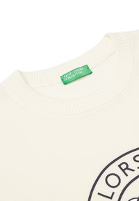 Crème-kleurige trui met een ribbelhals, voorzien van een opvallend rond logo in donker marineblauw. Merklabel in het groen aan de hals.