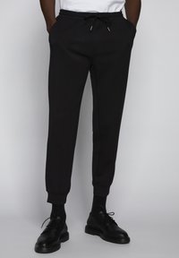 Pantalones de chándal negros en un tejido suave, con una cintura elástica ajustable, piernas entalladas y puños acanalados. Combinados con zapatos negros.
