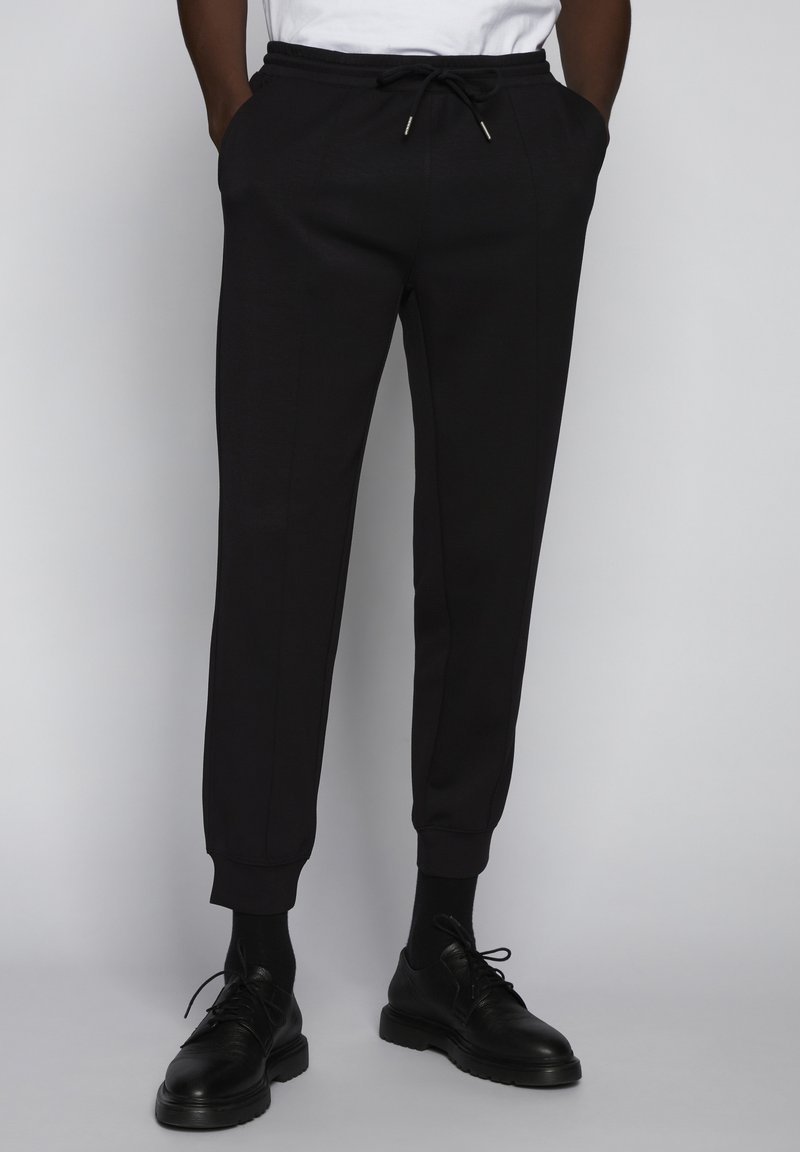 Pantalones de chándal negros en un tejido suave, con una cintura elástica ajustable, piernas entalladas y puños acanalados. Combinados con zapatos negros.