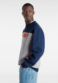 Marinblå och grå sweatshirt med röd logotyp, rundad halsringning och långa ärmar. Tyget är slätt och har kontrasterande färgpaneler.