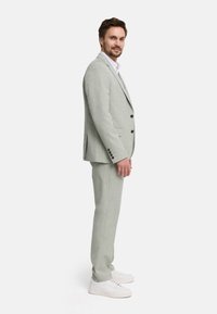 Man standing sideways wearing a light gray suit with black buttons, white shirt, and white sneakers against a plain background.