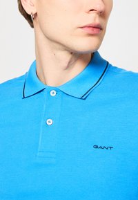 Tricou polo albastru dintr-un material texturat. Dispune de guler, două butoane și un logo brodat negru pe piept.