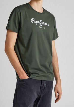 Man die een donker groen Pepe Jeans London T-shirt en donkerblauwe spijkerbroek draagt, staat met één hand in zijn zak tegen een effen achtergrond.