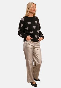 Maglione nero con texture soffice e motivi a cuore argentati, abbinato a pantaloni larghi argentati lucidi. La modella è in piedi con le mani nelle tasche.