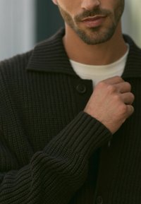 Cardigan en maille sombre avec un col évasé, texture côtelée, boutons sur le devant et manches longues. T-shirt blanc à col ras du cou visible en dessous.