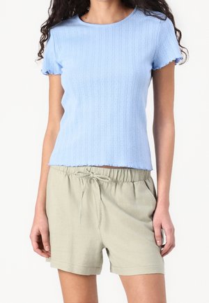 Kobieta w jasnoniebieskim prążkowanym topie z krótkim rękawem i beżowych szortach z troczkami, stoi z rozluźnionymi rękami na białym tle.