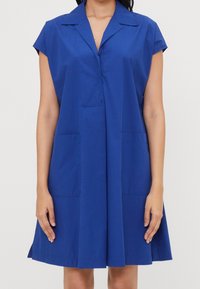 Donna che indossa un vestito senza maniche, di lunghezza al ginocchio, di un blu royal, con colletto e due tasche frontali, in piedi con le braccia rilassate ai fianchi.