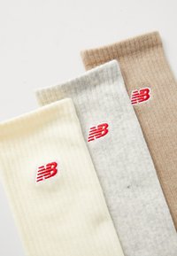 Tre paia di calzini a coste in crema, grigio e beige, ognuno con un logo "NB" rosso ricamato vicino alla parte superiore. Materiale morbido e tessile.