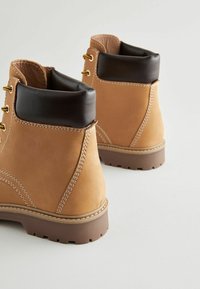 Botines de cuero marrón con un cuello acolchado de un marrón oscuro, cordones amarillos y una suela de goma resistente con patrones de huella visibles.