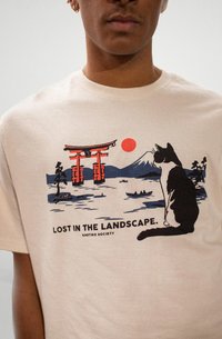 Person wearing a beige shirt with a graphic of a black and white cat looking at a red torii gate, sun, boats, and mountain, text "Lost in the landscape."