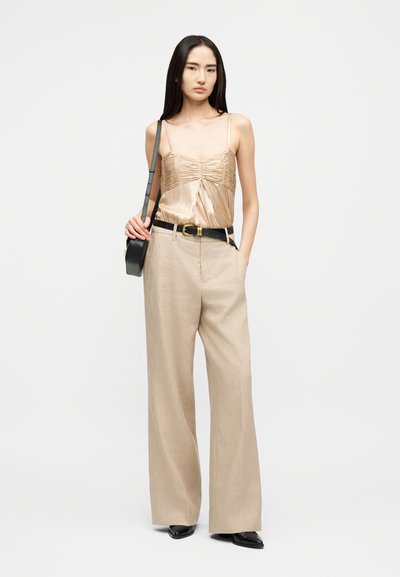 Γυναίκα που φοράει μπεζ σατέν κορμάκι με ραφές, μπεζ παντελόνι με φαρδιά μπατζάκια και μαύρη ζώνη, μαύρα μυτερά παπούτσια και κρατάει μαύρη τσάντα χιαστί.