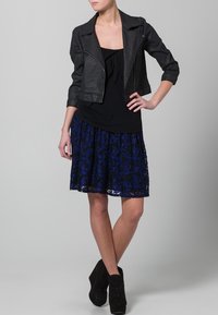 Chaqueta negra de cuero sintético corta con mangas tres cuartos, combinada con una camiseta de tirantes negra y una falda azul con patrón de encaje, de longitud por encima de la rodilla.