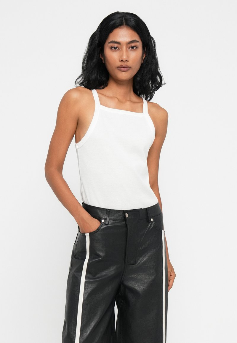 Top sin mangas blanco con escote cuadrado y ribete en contraste, combinado con pantalones de cuero negros de talle alto con franjas laterales.