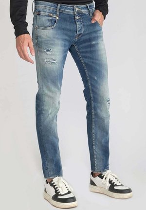 Mężczyzna w wyblakłych, podartych niebieskich dżinsach i czarno-białych trampkach, stojący z jedną ręką w kieszeni na jednolitym tle.