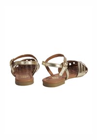 Metallisch goldene Sandalen mit offenem Riemendesign, flacher Sohle und verstellbarem Knöchelverschluss. Fußbett mit Holzstruktur und Aussparungen.