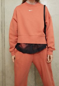 Korallfärgad sweatshirt med vit logotyp, oversized design, rundad halsringning och ribbade ärmslut, lagd över en svart genomskinlig skjorta och matchande byxor.