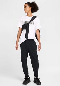 T-shirt blanche avec un graphique "AIR" en noir et gris, assortie à un pantalon de jogging noir avec un logo, et des baskets noires texturées.
