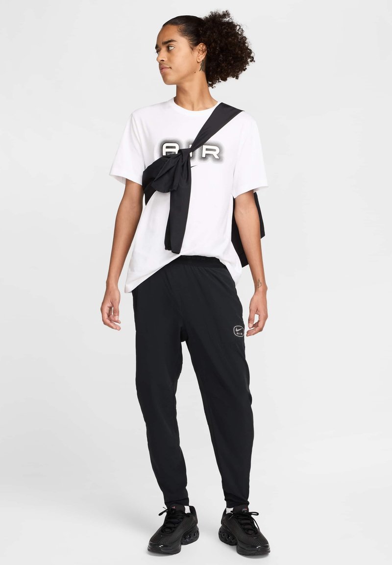 T-shirt blanche avec un graphique "AIR" en noir et gris, assortie à un pantalon de jogging noir avec un logo, et des baskets noires texturées.