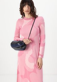 Rochie tricotată roz cu modele florale abstracte, mâneci lungi, decolteu rotund. Posetă de umăr neagră și bleumarin ținută într-o mână.