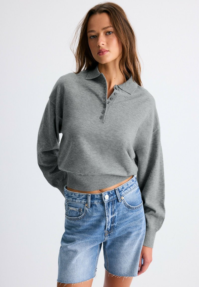 Jeune femme portant un polo gris à manches longues court et un short en jean bleu taille haute sur un fond uni.