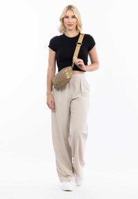 Borsa a tracolla metallica dorata con texture liscia, tracolla regolabile e chiusura con zip, indossata sopra una maglietta nera e pantaloni beigi.