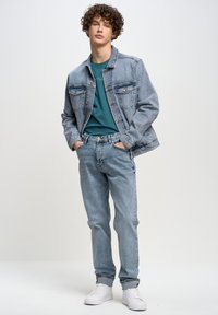 Chaqueta de mezclilla azul clara con dos bolsillos en el pecho, combinada con una camiseta verde azulado y jeans azul claro. Completada con zapatillas blancas.