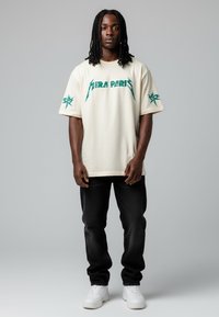 T-shirt crème à manches courtes avec un graphique vert du logo "AIRA PARIS", associé à un jean noir et des baskets blanches, se tenant devant un fond gris.