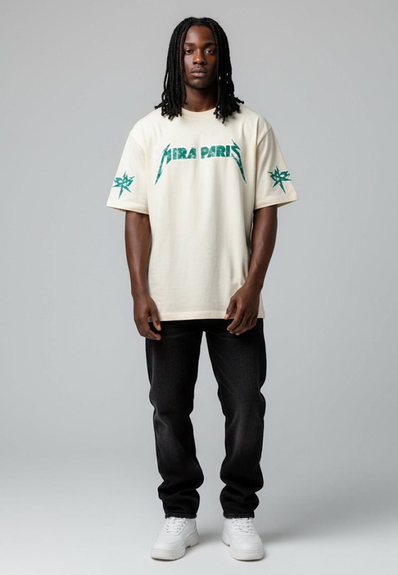 T-shirt crème à manches courtes avec un graphique vert du logo "AIRA PARIS", associé à un jean noir et des baskets blanches, se tenant devant un fond gris.