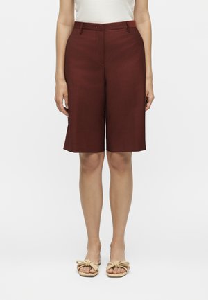 Žena nosiaca tmavé červené šortky na mieru dĺžky po kolená, biely nátelník bez rukávov a béžové sandále s detailmi z lana, stojaca na bielom pozadí.