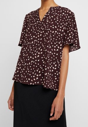 Vrouw draagt een korte mouw, losvallende bruine blouse met onregelmatige lichtbeige vlekken en een V-hals, gecombineerd met een zwarte rok.