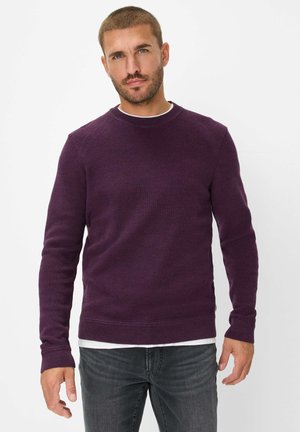 Leichtes lila Strickpullover mit langen Ärmeln, rundem Ausschnitt und gerippten Bündchen. Getragen über einem weißen Unterhemd, kombiniert mit dunkelgrauen Jeans.