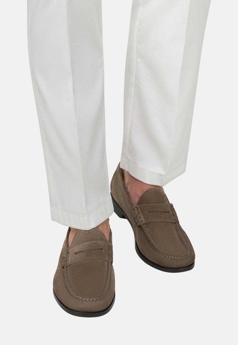 Mocassins en daim marron portés avec un pantalon blanc tailleur, vus de mi-mollet vers le bas sur un fond uni.