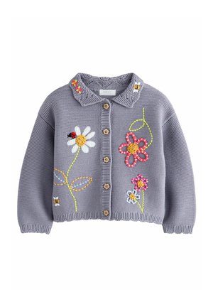 Enfant portant un cardigan violet boutonné avec des fleurs brodées, un jean bleu à jambes larges et des baskets roses, debout sur un fond neutre.