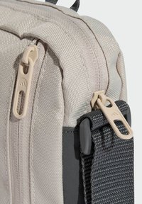 Beżowa torba materiałowa o teksturowanej powierzchni, wyposażona w gładki zamek błyskawiczny, beżowe suwaki i czarny pasek z plecionym wzorem.