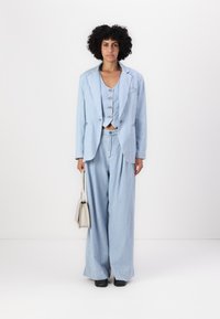 Completo azzurro chiaro a righe con gilet corto, blazer oversize e pantaloni a gamba larga. Presenta bottoni argentati e una borsa beige.