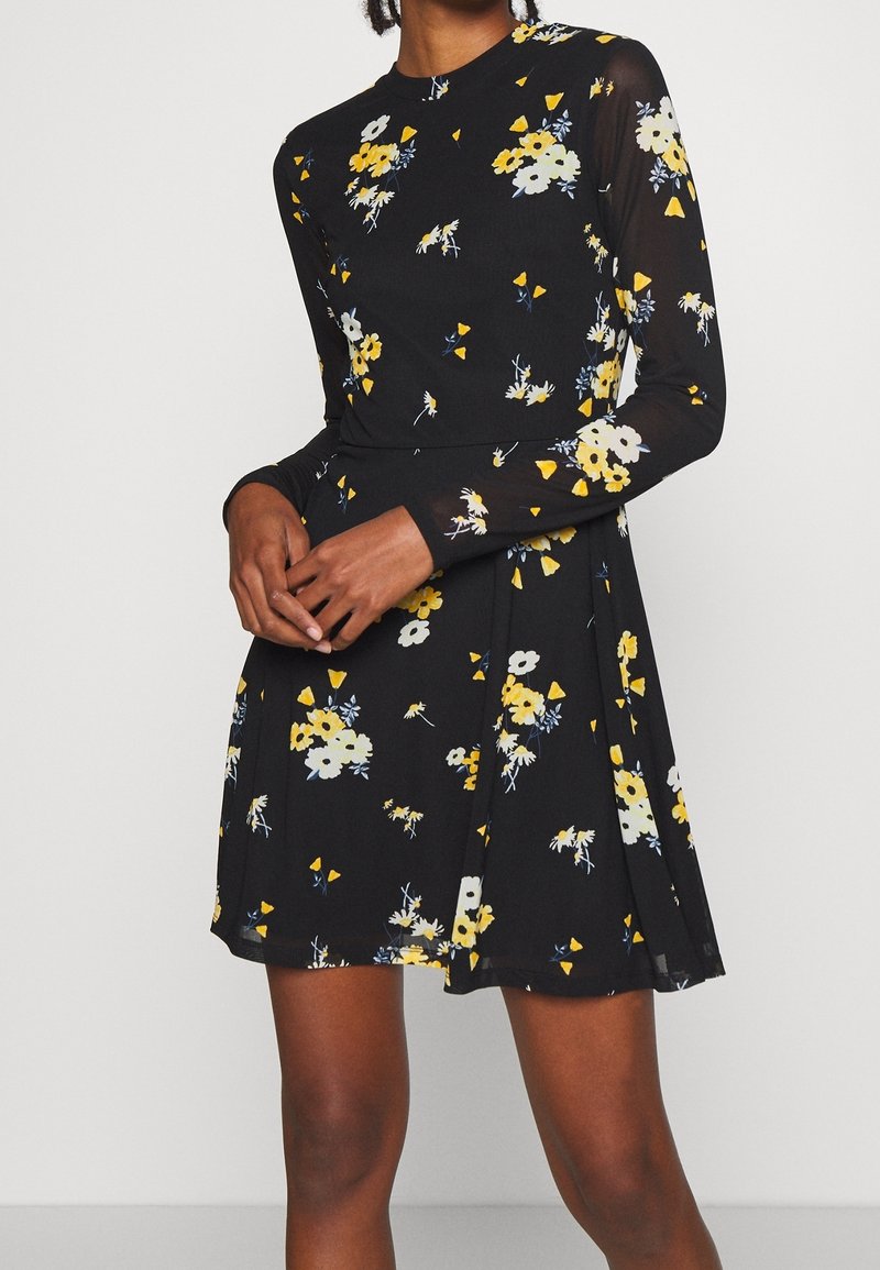 Femme portant une robe noire à manches longues avec un imprimé floral jaune, blanc et bleu, debout avec les mains doucement jointes devant.