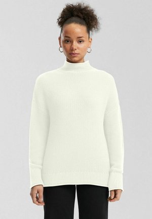 Kobieta z kręconymi włosami związanymi w wysoki kucyk, nosząca złote kolczyki koła, biały dziany sweter z golfem oraz czarne spodnie, stojąca w neutralnej pozie.