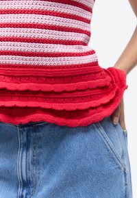 Close-up van een gelaagde rode en roze gestreepte gebreide top gecombineerd met blauwe spijkerbroek, met een hand rustend op de heup.