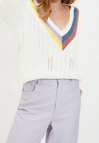 Pull en maille torsadée blanche avec un col en V orné d'accents rayés multicolores en violet, jaune et rouge. Assorti à un pantalon lavande clair.