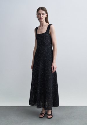 Nő áll egy ujjatlan, fekete, texturált maxi ruhában és fekete pántos szandálban, sima világosszürke háttér előtt.