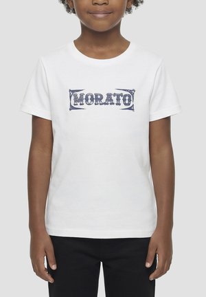 Niño con camiseta blanca de manga corta con texto azul "MORATO", combinado con pantalones negros, de pie sobre un fondo claro y liso.