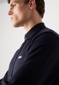 Mørkeblå polo-skjorte med en myk tekstur, med knapper i kragen og et subtilt logo på brystet. Nærbilde av sideprofilen.