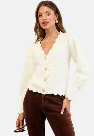 Donna che indossa un cardigan bianco con bordo a scallop e bottoni decorativi e pantaloni di velluto a coste marroni, mentre tiene occhiali tartarugati in una mano.