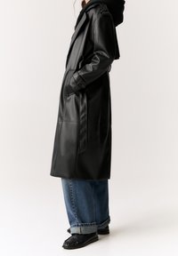 Trench-coat en cuir noir avec une taille cintrée par une ceinture, une coupe ajustée et des coutures détaillées. Associé à un jean bleu délavé, ample et avec revers.