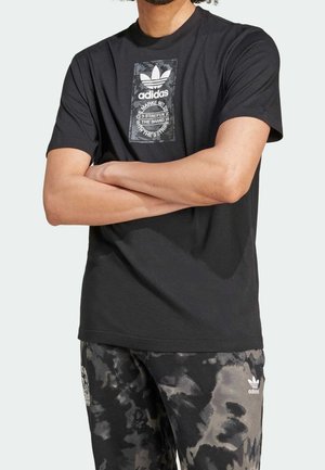 Black cotton t-shirt featuring a large grey Adidas logo on a rectangular patch. Paired with grey and black tie-dye patterned pants.