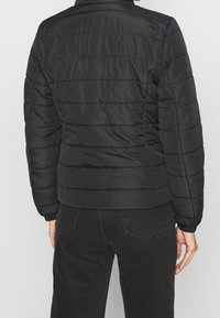 Chaqueta negra acolchada con cuello alto, que presenta una textura suave y puños acanalados. La vista trasera muestra costuras horizontales y un corte entallado.